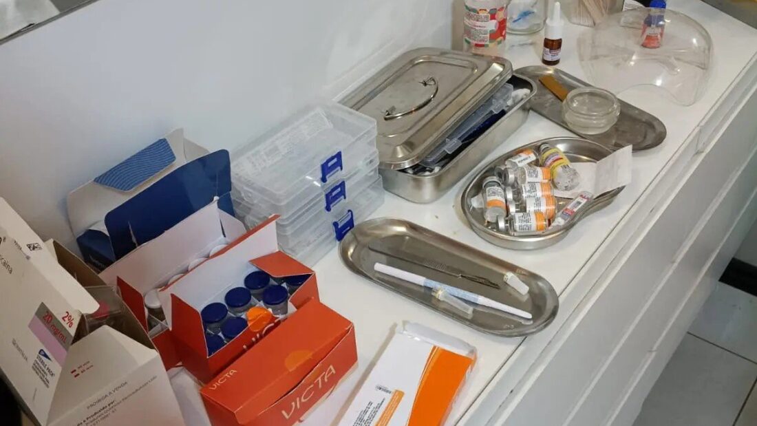 Bancada branca com caixas de medicamentos, frascos e bandejas metálicas com instrumentos dentro de uma sala de procedimentos