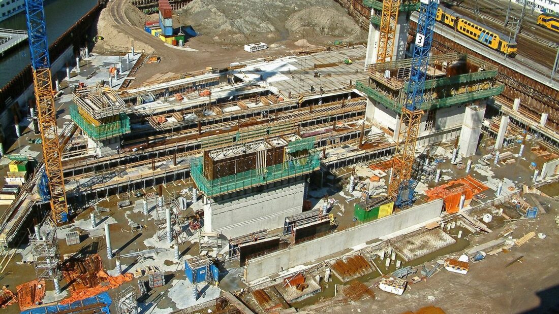 Vista do alto de um grande canteiro de obras, com estruturas de concreto em execução, guindastes amarelos e escavações