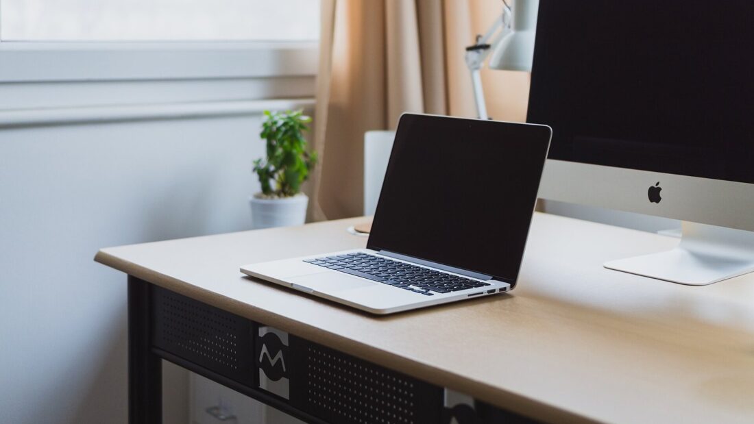 Notebook aberto sobre uma mesa de escritório branca, ao lado de um monitor de computador e de um vaso com planta