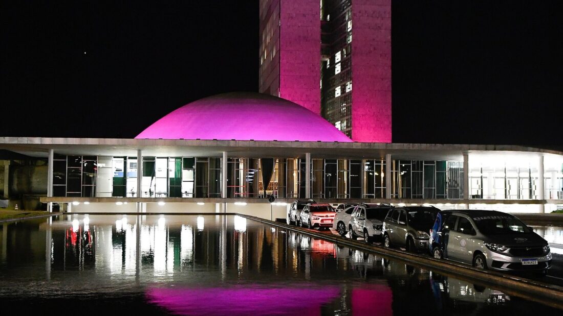 Cúpula do Senado Federal e prédio do Congresso Nacional iluminados de roxo à noite, refletidos no espelho d'água