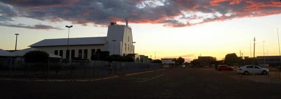 Fim de tarde em Mozarlândia, com a silhueta da torre da Igreja Matriz e de construções baixas sob céu alaranjado