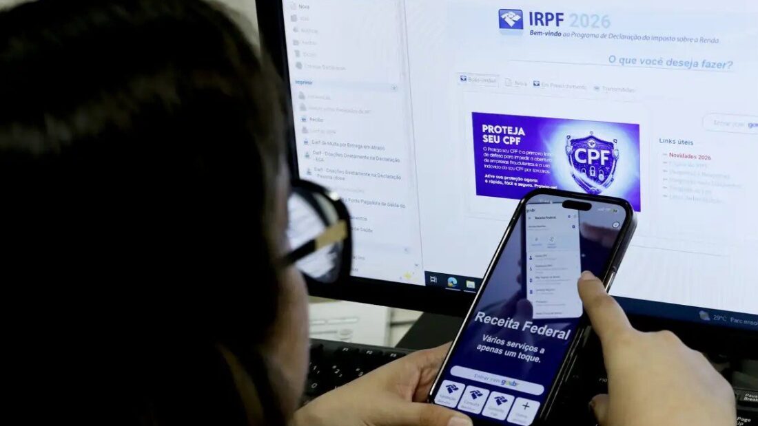 Pessoa consulta o aplicativo da Receita Federal no celular diante de um computador aberto na página do Imposto de Renda 2026