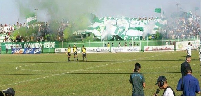 Torcida do Esporte Clube Rio Verde com bandeiras verdes e brancas e sinalizadores de fumaça verde na arquibancada, com jogadores e árbitros no gramado