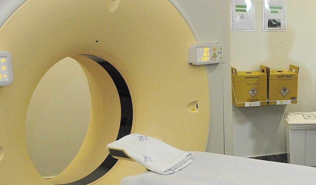 Aparelho de tomografia em uma sala de exames de hospital, com a maca coberta por lençol branco