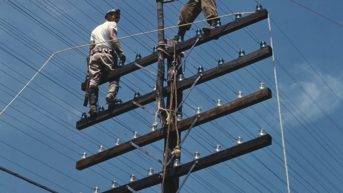 Dois eletricistas trabalham no alto de um poste de madeira com várias cruzetas e fios, contra o céu azul