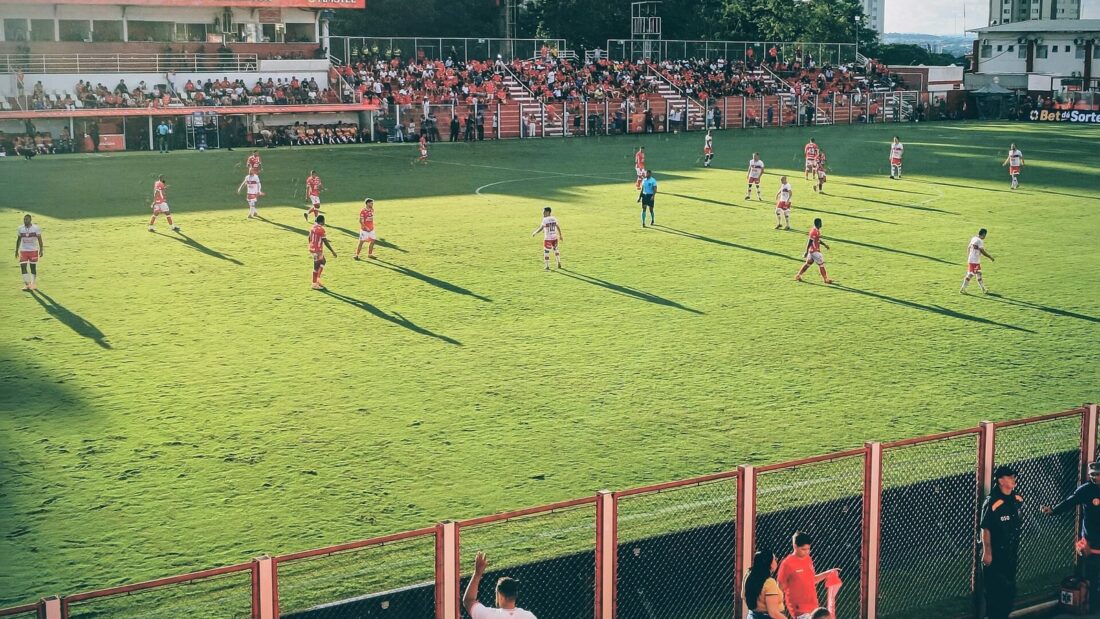 Vista da arquibancada para o gramado do Estádio Onésio Brasileiro Alvarenga durante uma partida, com torcedores de vermelho ao fundo e prédios atrás do estádio