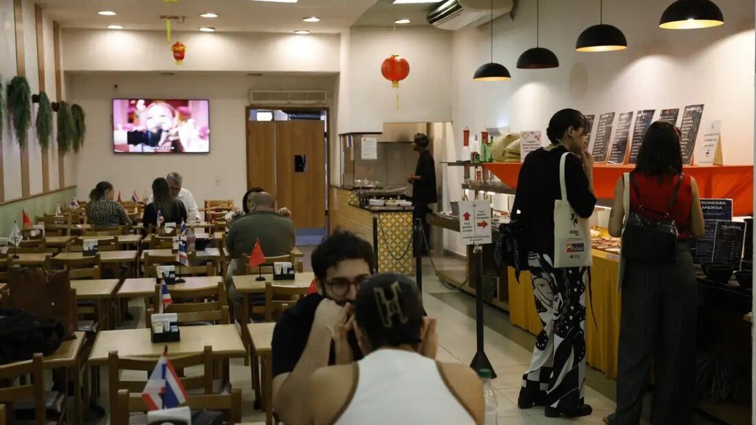 Salão de restaurante com mesas de madeira ocupadas por clientes, luminárias pendentes, televisão na parede e balcão de bufê ao fundo