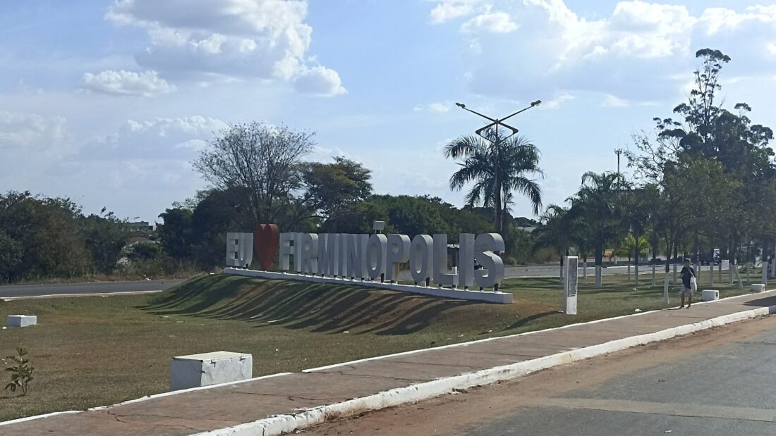 Letreiro com o nome Firminópolis num canteiro gramado à beira de uma avenida larga, com árvores e palmeiras ao fundo e céu com poucas nuvens