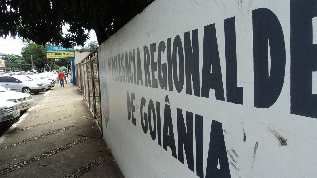 Muro branco com a inscrição em letras pretas Delegacia Regional de Goiânia, ao lado de uma calçada com carros estacionados