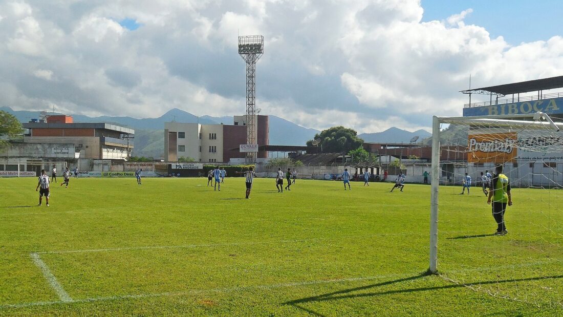 Partida de futebol em campo gramado de estádio pequeno, com jogadores, trave em primeiro plano e torre de iluminação ao fundo