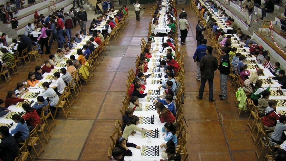 Ginásio com longas fileiras de mesas e tabuleiros de xadrez ocupadas por crianças e jovens durante um torneio
