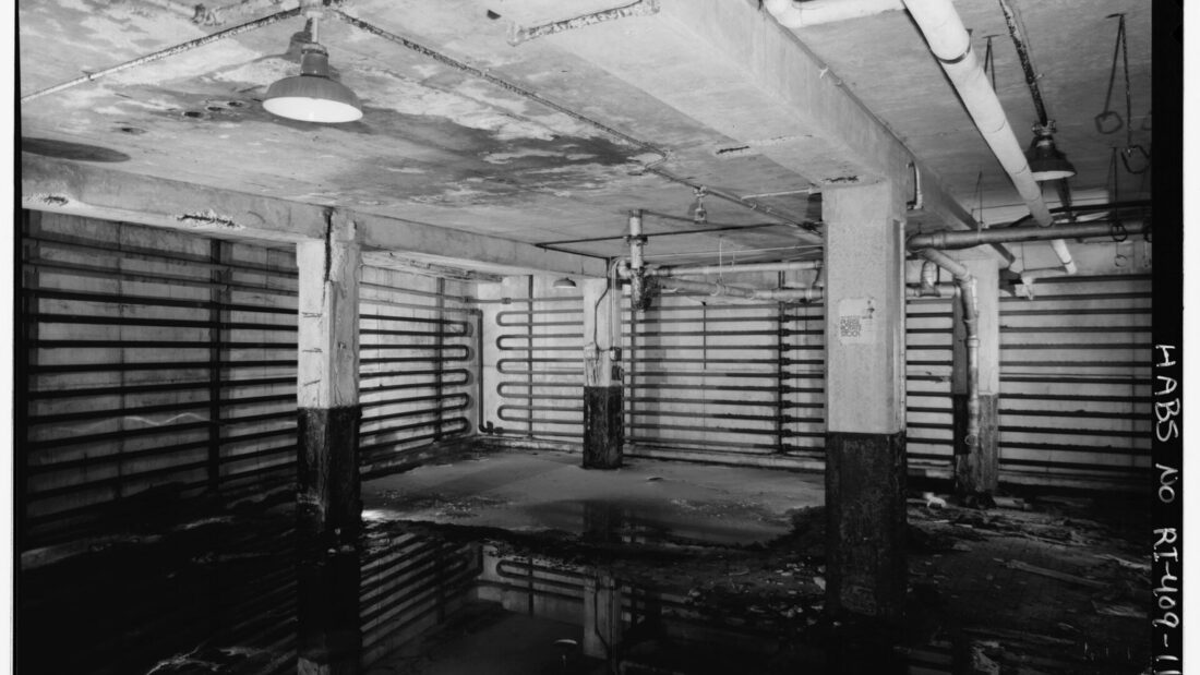 Foto em preto e branco do subsolo de um antigo frigorífico, com colunas de concreto e tubulações de refrigeração presas às paredes e ao teto