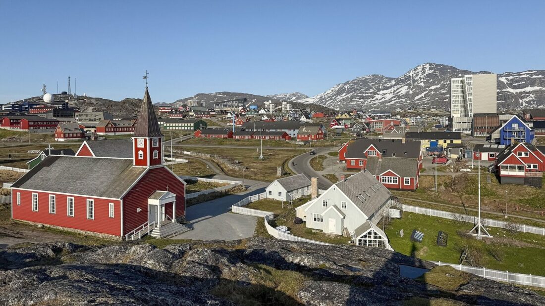 Vista de Nuuk, capital da Groenlândia, com a catedral de madeira vermelha em primeiro plano, casas coloridas, prédios e montanhas com neve ao fundo