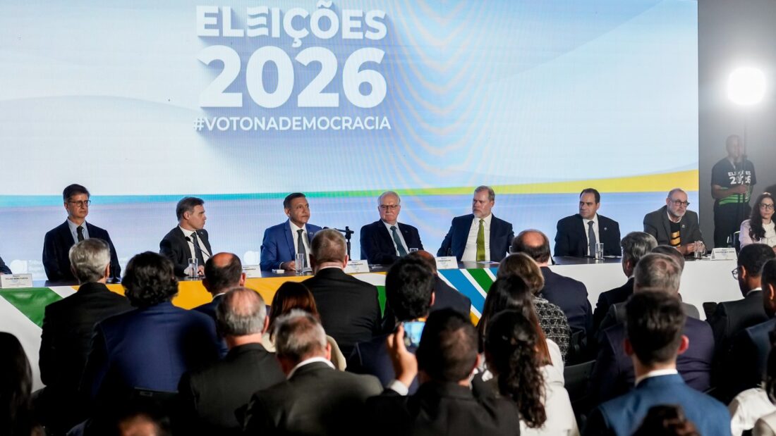 Ministros do TSE sentados à mesa durante entrevista coletiva, diante de painel das Eleições 2026 e de jornalistas