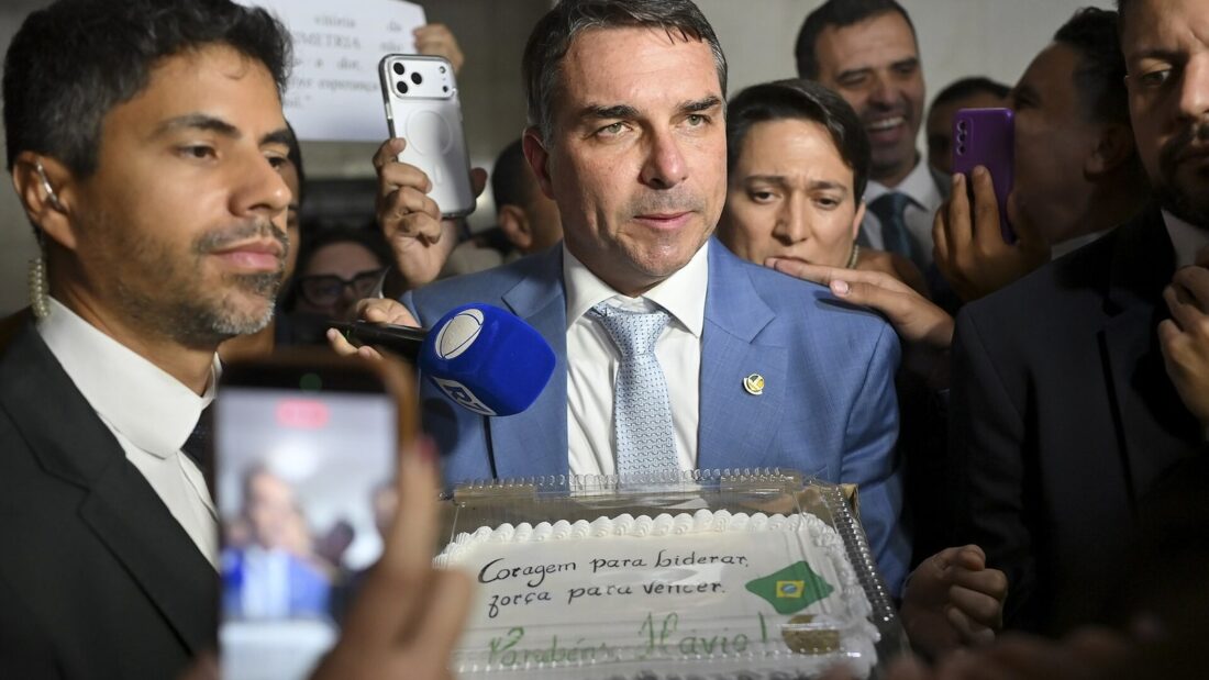 Flávio Bolsonaro, de terno azul, cercado por apoiadores e jornalistas com microfones e celulares, diante de um bolo decorado com uma frase de incentivo