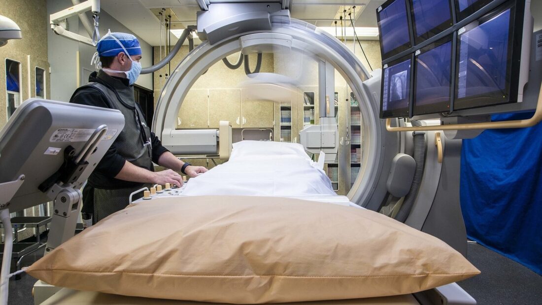 Sala de radiologia de hospital com aparelho de raio X em arco sobre uma maca, monitores no alto e um técnico de máscara e touca operando o equipamento