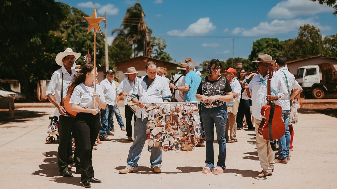 Foliões de Reis caminham em grupo por rua de terra de um distrito rural, com viola, bandeira enfeitada e chapéus, sob céu azul