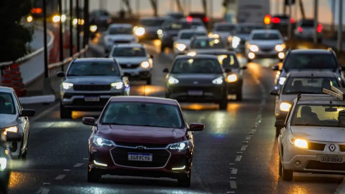 Fila de carros com faróis acesos em avenida congestionada no fim do dia