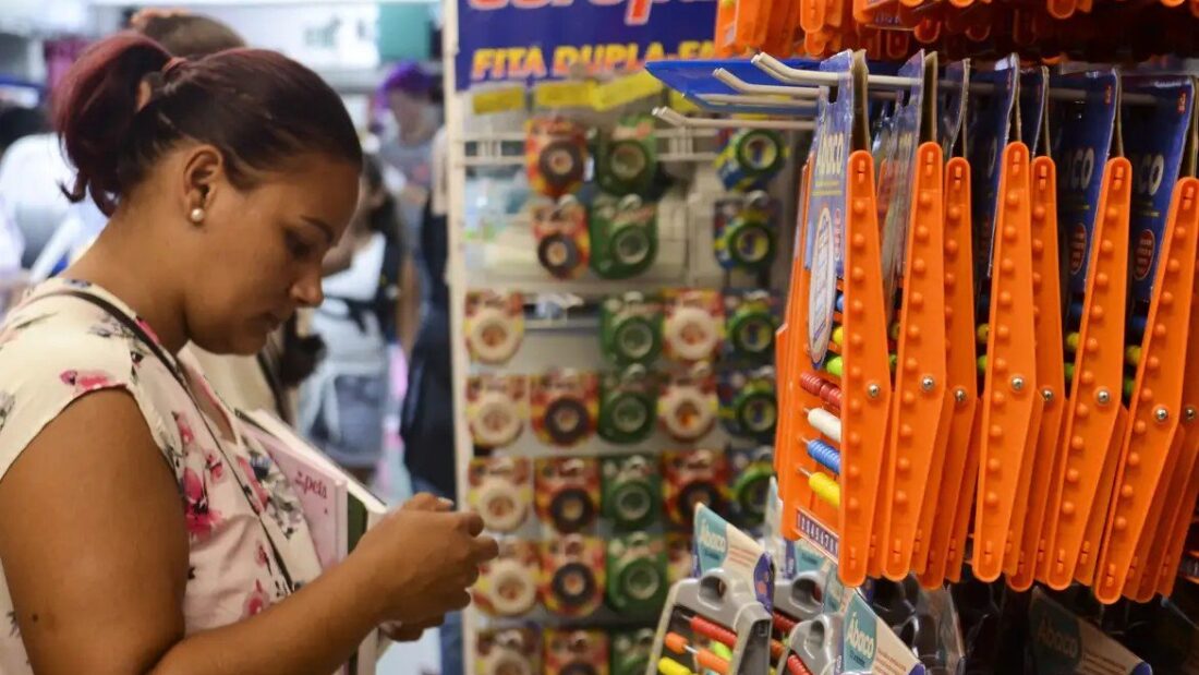 Mulher escolhe produtos em uma loja de material escolar, diante de um expositor com fitas, réguas e estojos coloridos pendurados