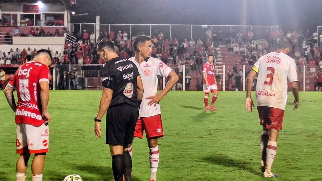 Jogadores de camisa vermelha do Vila Nova e o árbitro, de preto, no gramado do estádio Onésio Brasileiro Alvarenga, à noite, com torcida na arquibancada
