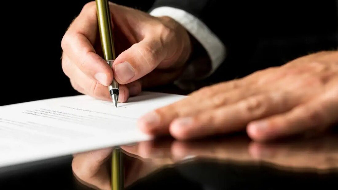 Mão segurando uma caneta assina um documento sobre uma mesa escura